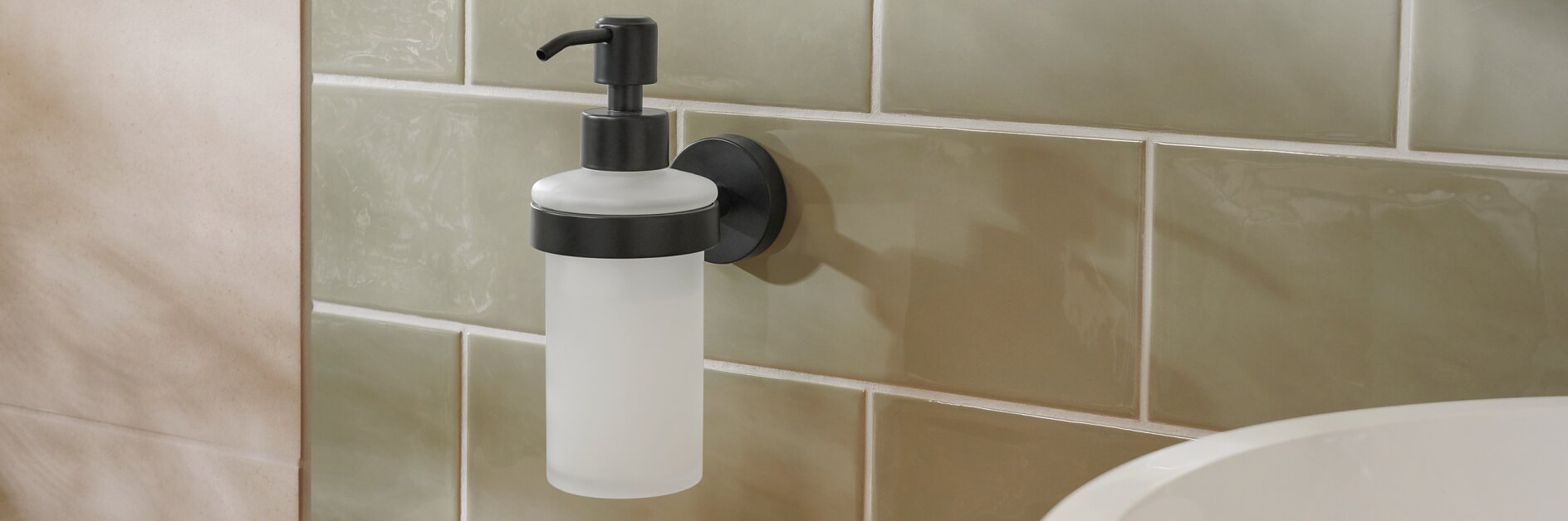 En väggmonterad tvålpump med en svart pump är fäst på en kaklad vägg med tesatejp. Kaklet är ljusgrönt och under det syns en bänkskiva i trä som delvis rymmer ett runt vitt handfat och ett rosa fat med guldsmycken.