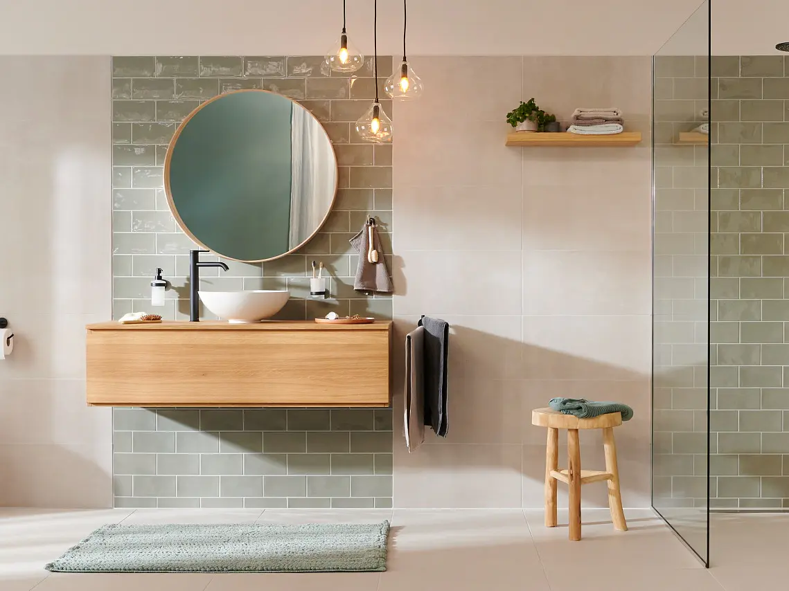 Ett modernt badrum har en vägghängd toalett och ett träskåp med en rund spegel. Duschområdet är inglasat med gröna kakelplattor. En handduk hänger på väggen med hjälp av tesa-tejp, och en liten träpall står nära en grön matta. Växter finns i närheten. (This text has been generated by AI)