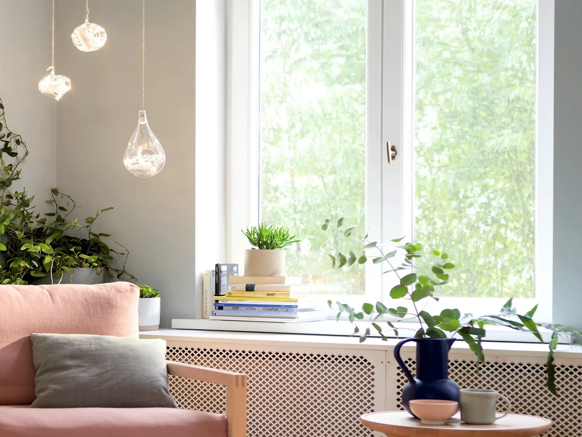 Ett ljust vardagsrum har en ljusrosa soffa med en grå kudde och kompletteras av ett litet bord med växter och en vas. Hängande pendellampor är placerade till vänster, medan solljus strömmar in genom ett stort fönster till höger och lyser upp böcker och fler växter på fönsterbrädan. (This text has been generated by AI)