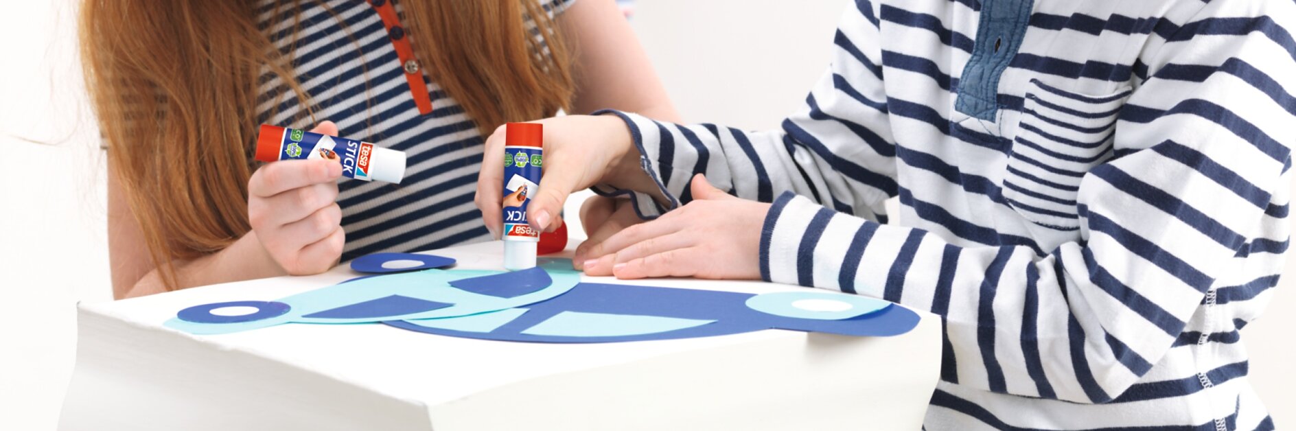 vå barn gör pyssel med papper och limstift på ett bord.