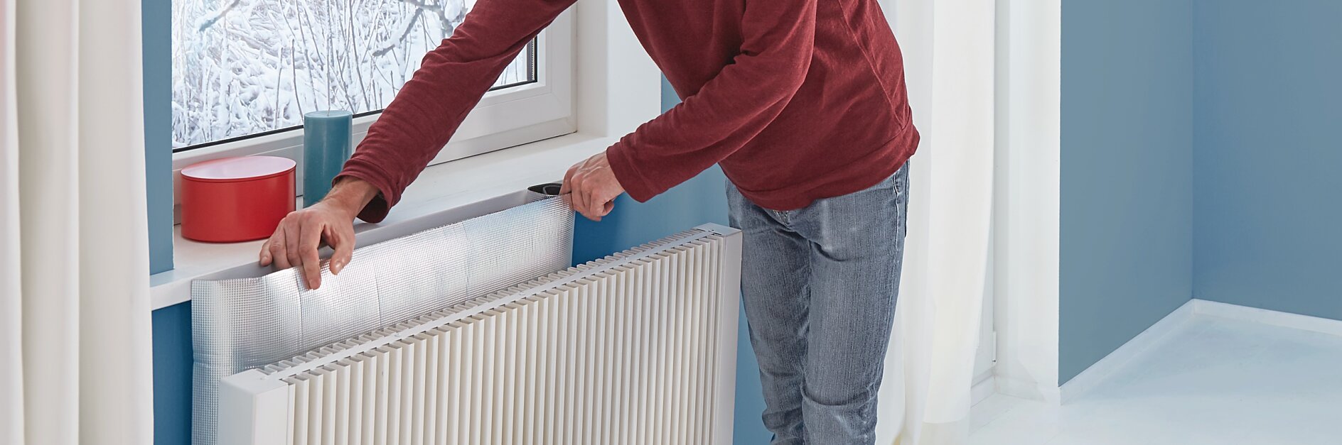 En man i röd tröja och jeans installerar folieisolering bakom ett element med tesa-tejp. En snöig utsikt syns genom fönstret, och rummet har blå väggar med vita gardiner. (This text has been generated by AI)