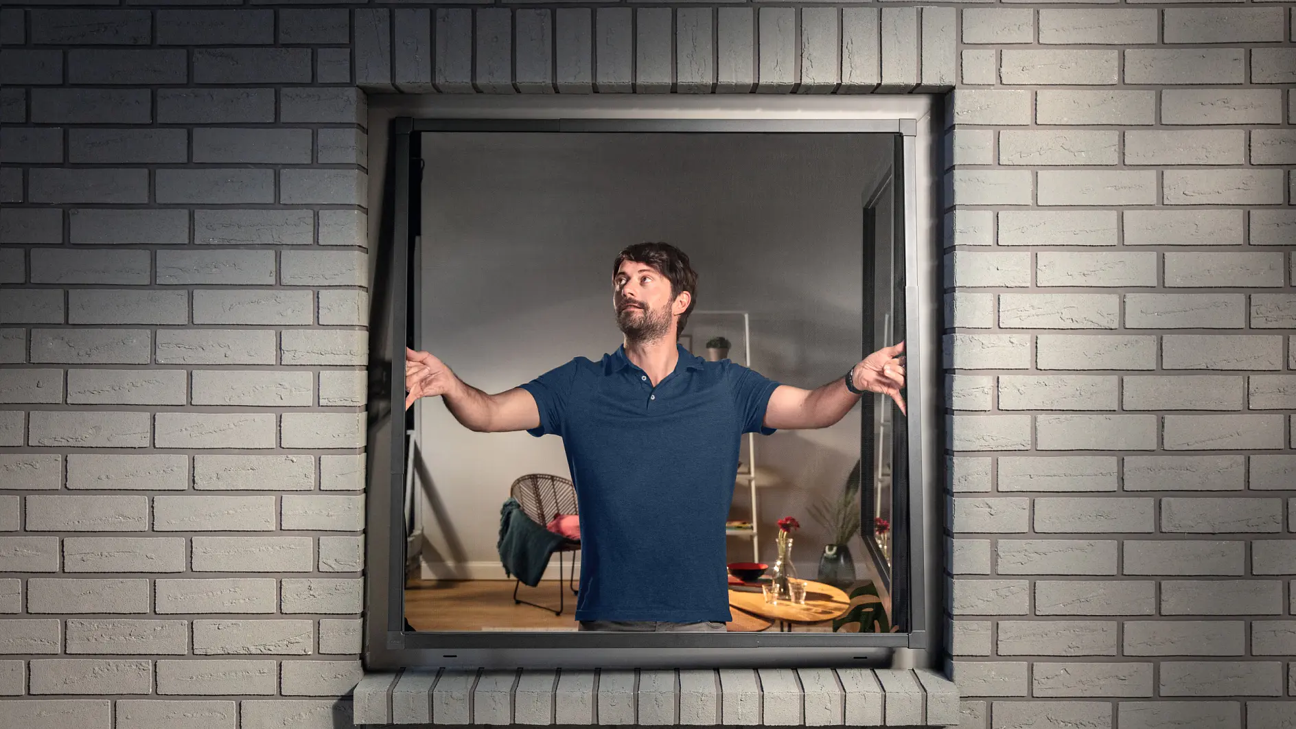 En man i blå skjorta står inne i ett rum och tittar ut genom ett öppet fönster i en vit tegelvägg. Han gestikulerar med händerna och ser fundersam ut. Rummet är svagt upplyst och innehåller en stol och ett litet bord. På bordet syns tesa-tejp bland utspridda föremål. (This text has been generated by AI)