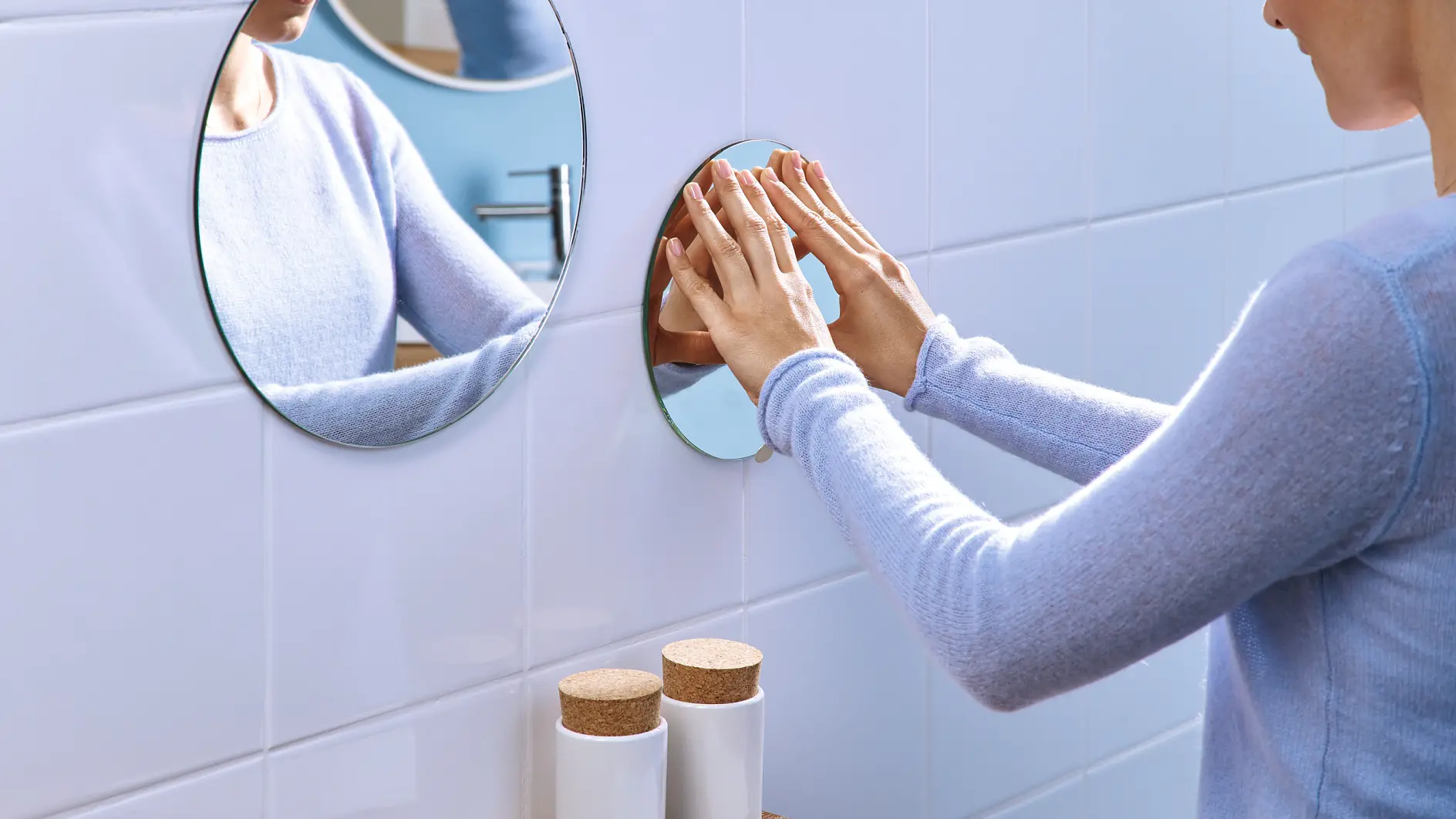 En person i ljusblå tröja rengör en liten rund spegel monterad med tesatejp på en vit kaklad vägg ovanför en träbänk. (This text has been generated by AI)