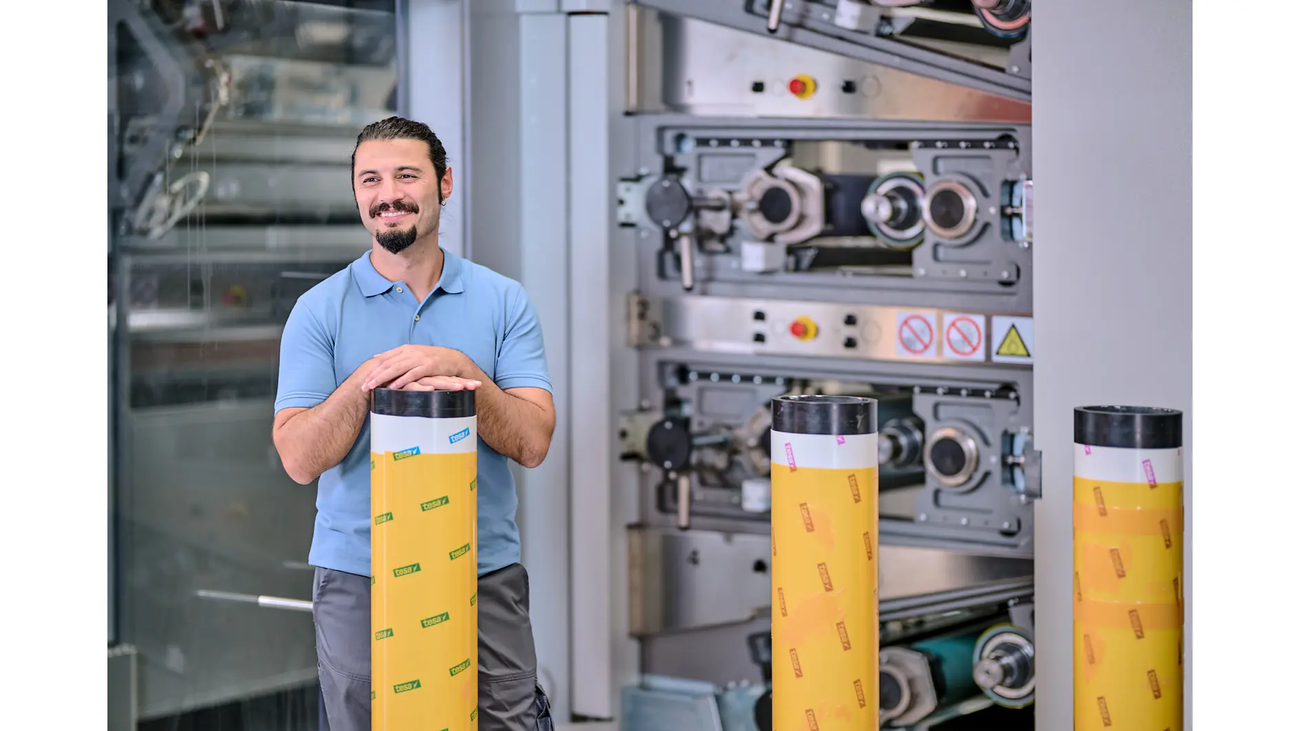 En man med skägg och hästsvans, iklädd en blå pikétröja, står leende med händerna på ett stort cylindriskt föremål insvept i gul tesa-tejp. Industriella maskiner syns i bakgrunden. (This text has been generated by AI)