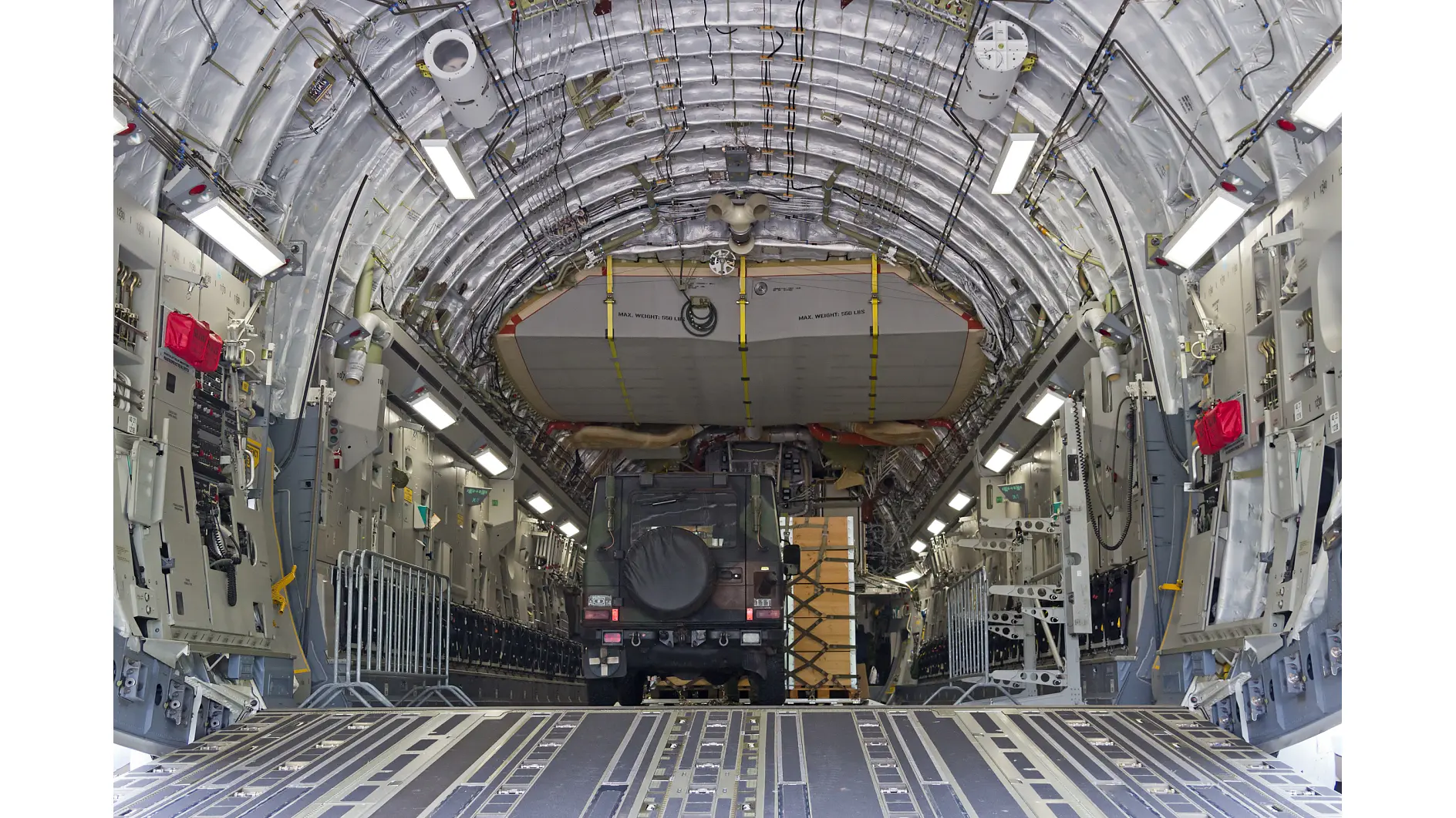 Interiörvy av ett stort militärt transportflygplan med rampen öppen, där ett fordon och last som är säkrade med tesa-tejp syns bland öppna paneler. (This text has been generated by AI)