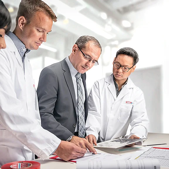 Four people are gathered around a table in a bright office. Two are wearing lab coats, one is in a suit, and one is partially visible. They are focused on documents laid out on the table, which include diagrams labeled with tesa tape. A laptop displaying pertinent data is open nearby. (This text has been generated by AI)