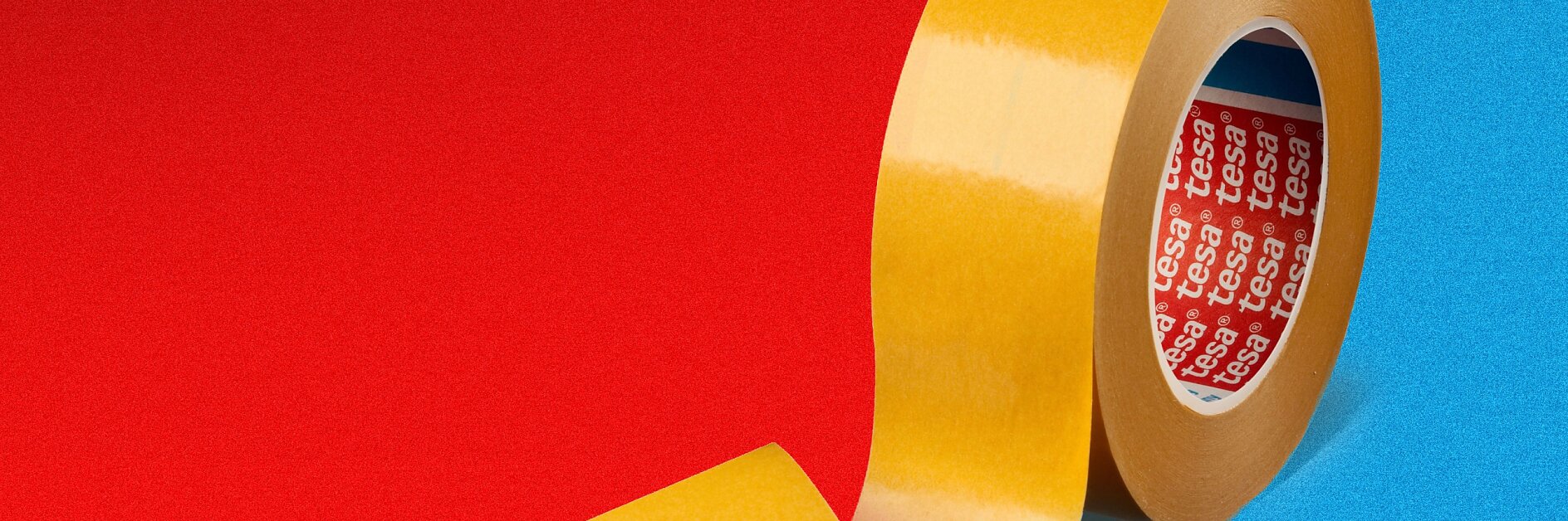 A roll of yellow tesa tape stands upright against a red and blue split background. The loose end of the tesa tape curls outward. The inside of the roll has a red label with white text.