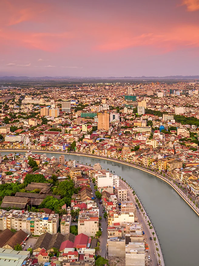 Aerial view of a cityscape during sunset with a river curving through the center. The skyline is dotted with buildings of varying heights. The sky displays hues of orange and pink, creating a warm ambiance. (This text has been generated by AI)