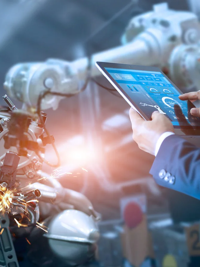 A person in a blue suit operates a tablet while overseeing a robotic assembly line. The robotics are working on an engine, with visible sparks flying. A futuristic interface is displayed on the tablet screen. (This text has been generated by AI)