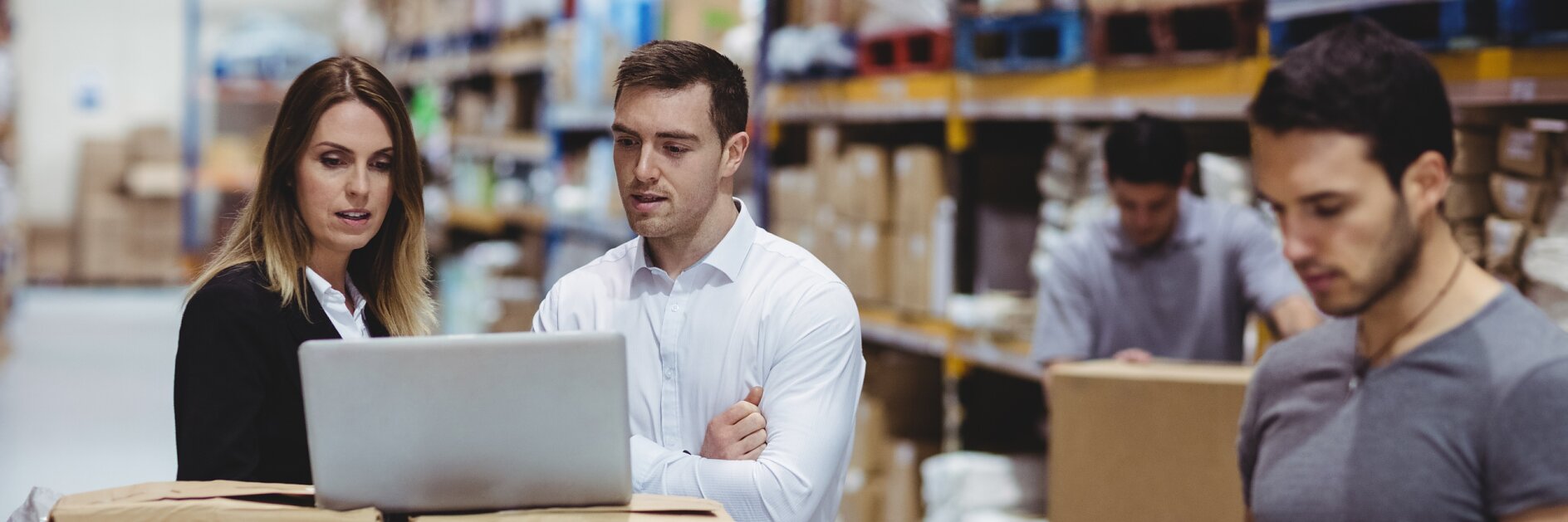 A group of warehouse workers stand by a table filled with packages. Two are engaged with a laptop, while another tallies items using a handheld device. Shelves stocked with boxes and supplies, including tesa tape, are visible in the background.