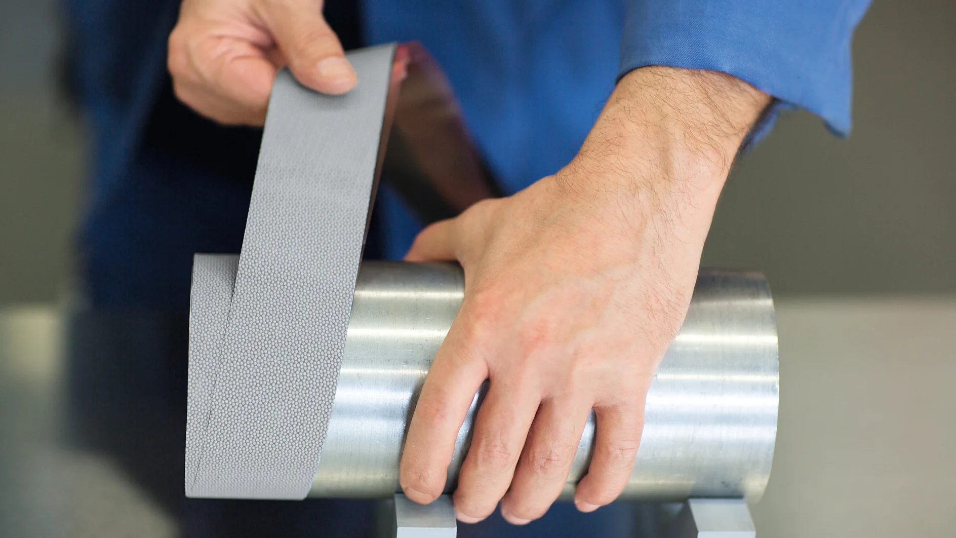 A person in a blue shirt demonstrates applying a strip of metallic, textured tesa tape to a cylindrical steel object. The object is placed on two gray stands on a smooth surface. (This text has been generated by AI)