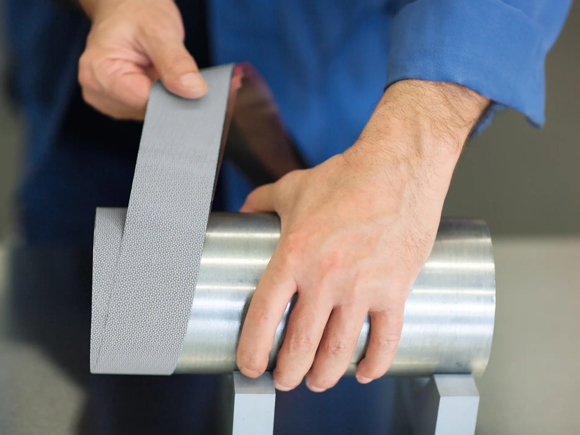 A person in a blue shirt demonstrates applying a strip of metallic, textured tesa tape to a cylindrical steel object. The object is placed on two gray stands on a smooth surface.