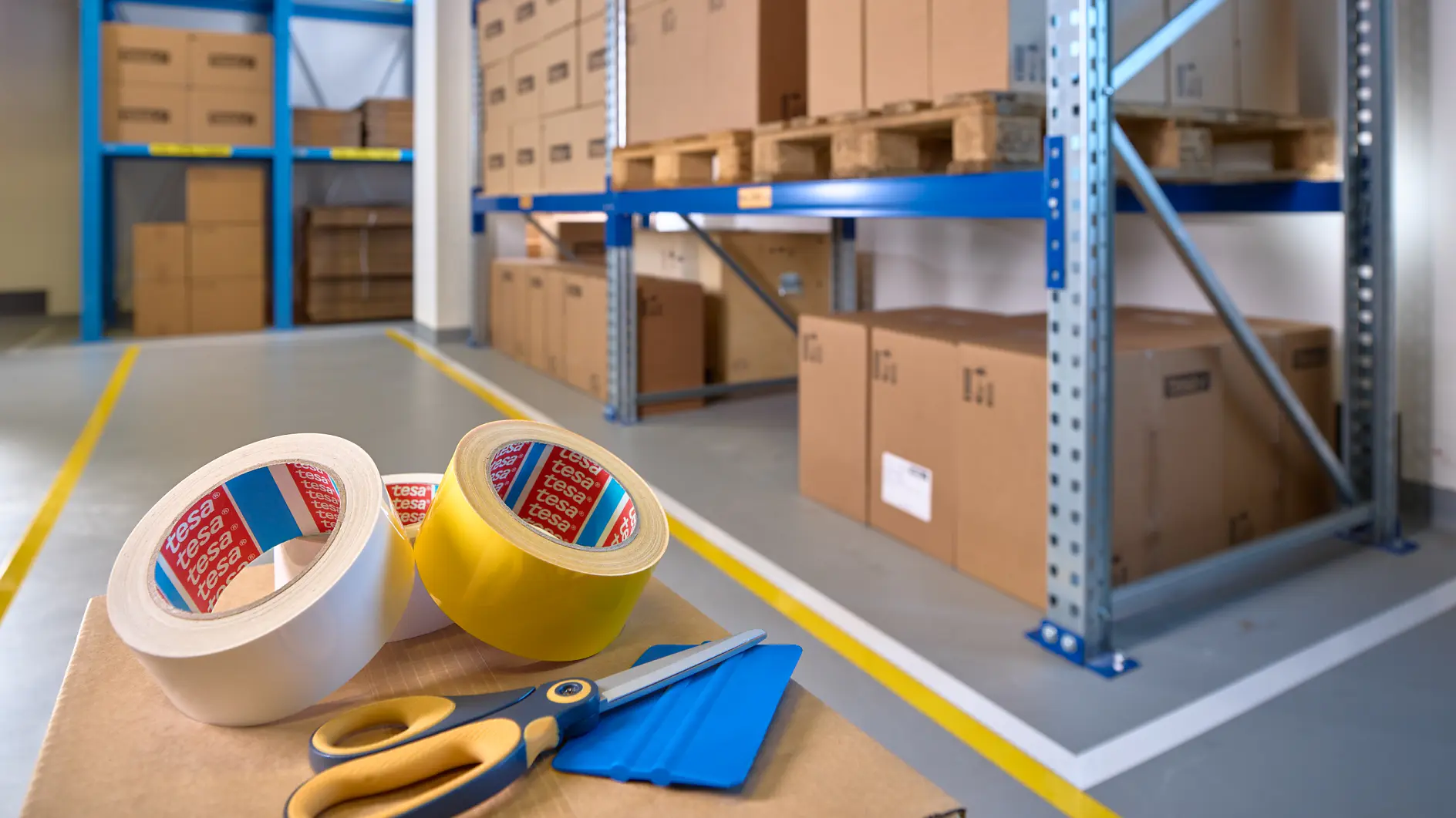 A warehouse interior with shelves stacked with cardboard boxes on wooden pallets. In the foreground, there are rolls of tesa tape, a pair of scissors, and a box cutter on a table. The floor is marked with yellow lines.