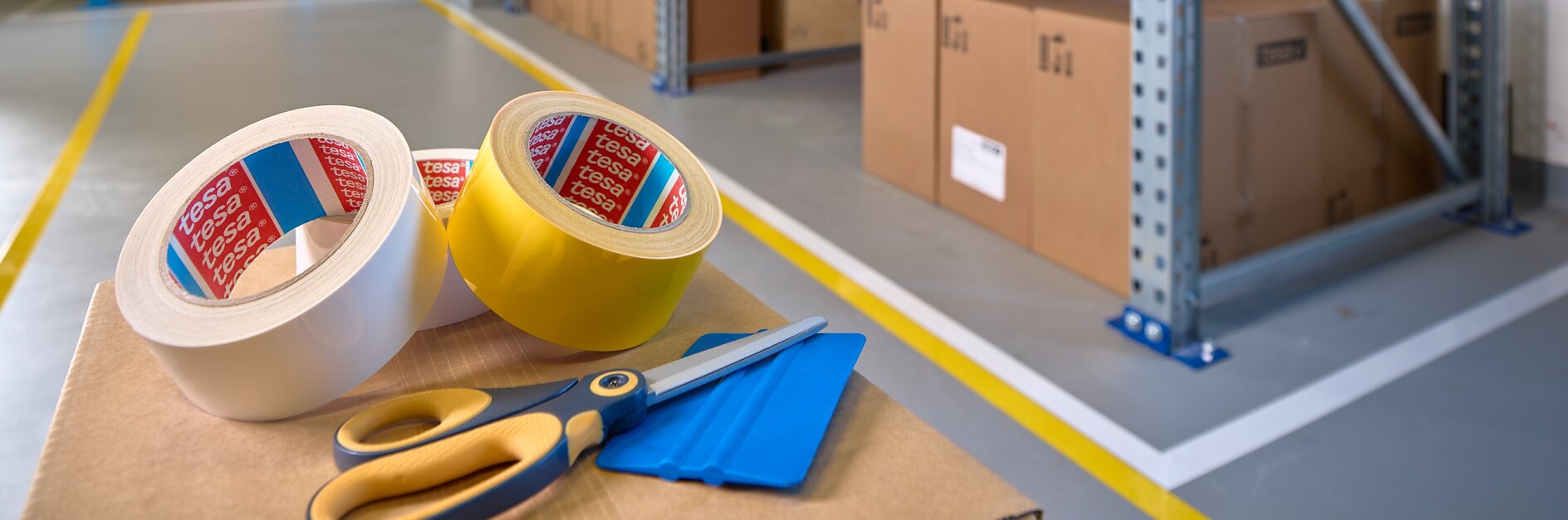 A warehouse interior with shelves stacked with cardboard boxes on wooden pallets. In the foreground, there are rolls of tesa tape, a pair of scissors, and a box cutter on a table. The floor is marked with yellow lines.