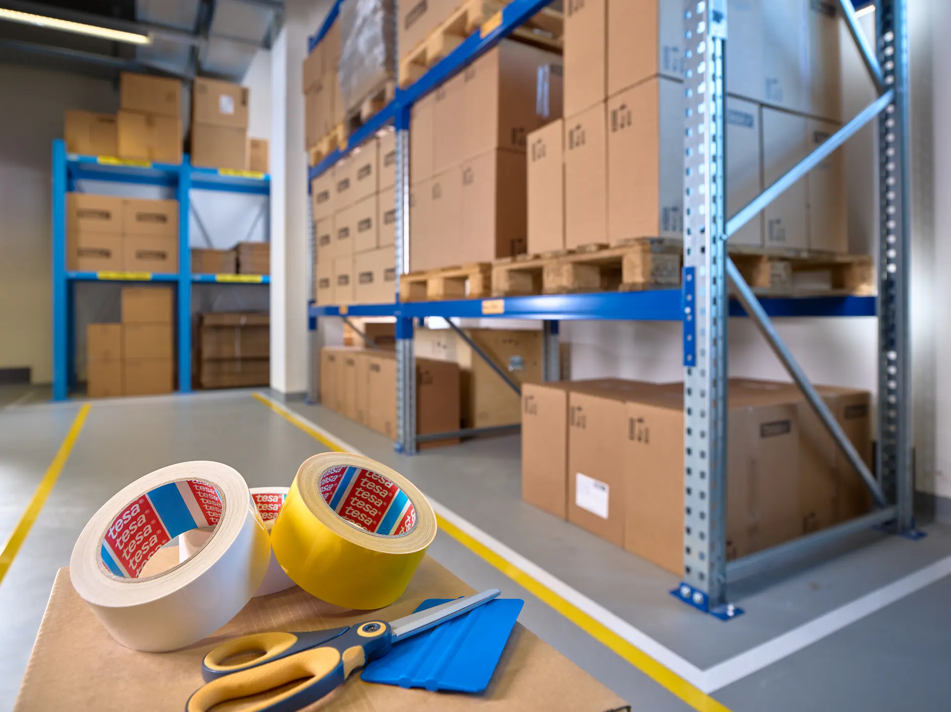 A warehouse interior with shelves stacked with cardboard boxes on wooden pallets. In the foreground, there are rolls of tesa tape, a pair of scissors, and a box cutter on a table. The floor is marked with yellow lines.