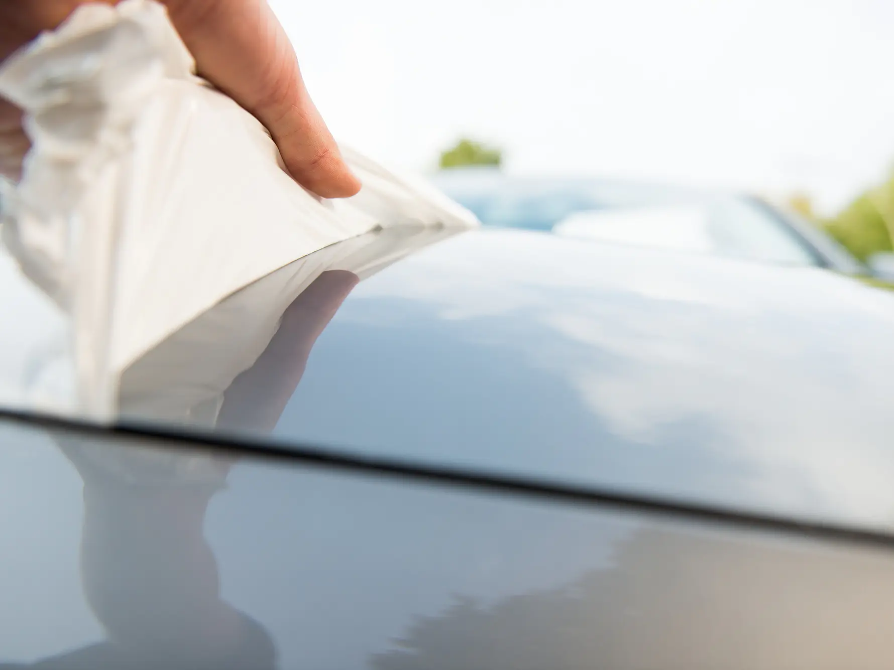 A hand is applying or removing a white protective film from the surface of a silver car using tesa tape. The close-up shot captures the action with a blurred background of greenery and other cars.