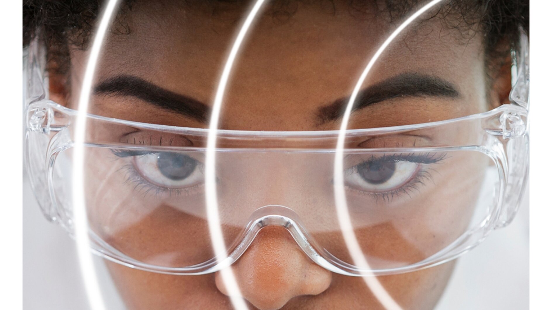A person wearing clear safety goggles is seen in close-up, with their face partially obscured by concentric circular white lines made of tesa tape. They have short curly hair and a focused expression. The background is white, and the image has a futuristic feel. (This text has been generated by AI)