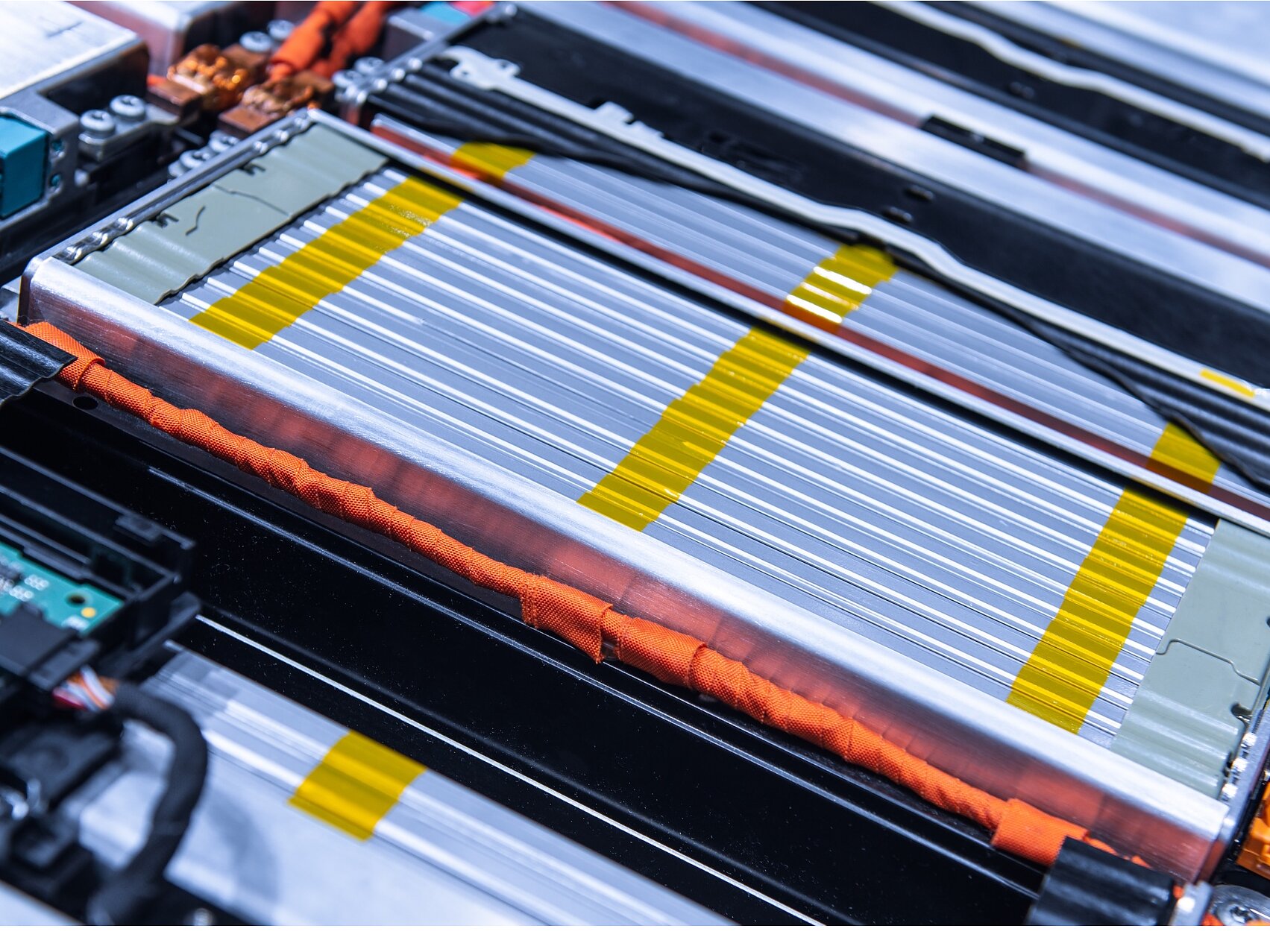 Close-up of a complex electrical circuit board and battery pack, featuring silver and yellow striped battery cells, orange wires, and a green circuit board. The components are tightly arranged within a metallic frame using tesa tape. (This text has been generated by AI)
