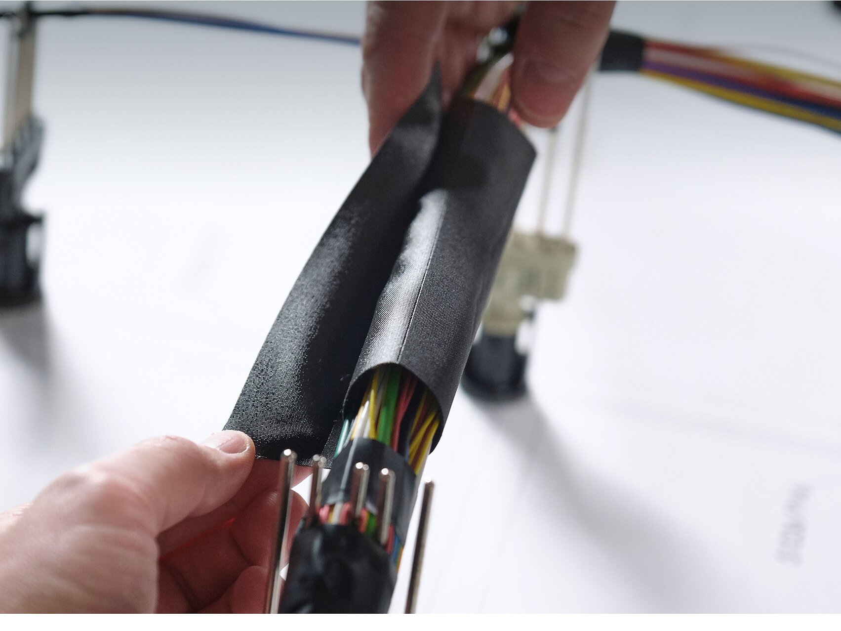 Close-up of hands wrapping a bundle of multicolored electrical wires with black insulating tesa tape. The wires are organized and connected to plastic connectors. A blurred background shows more wires and a surface with printed text. (This text has been generated by AI)