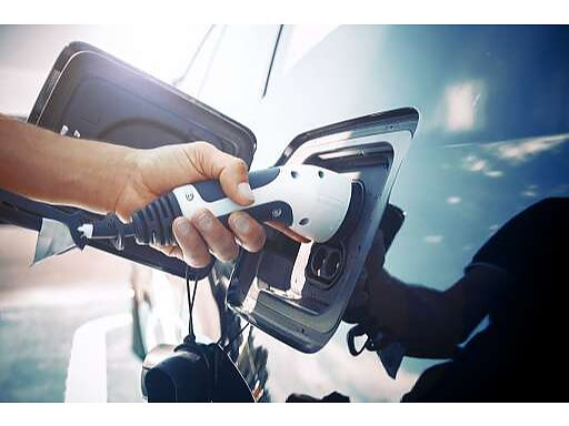 A person connects a charging cable to the charging port of an electric vehicle. The image is captured from a close-up angle, highlighting the hand gripping the cable against the cars shiny blue surface. (This text has been generated by AI)