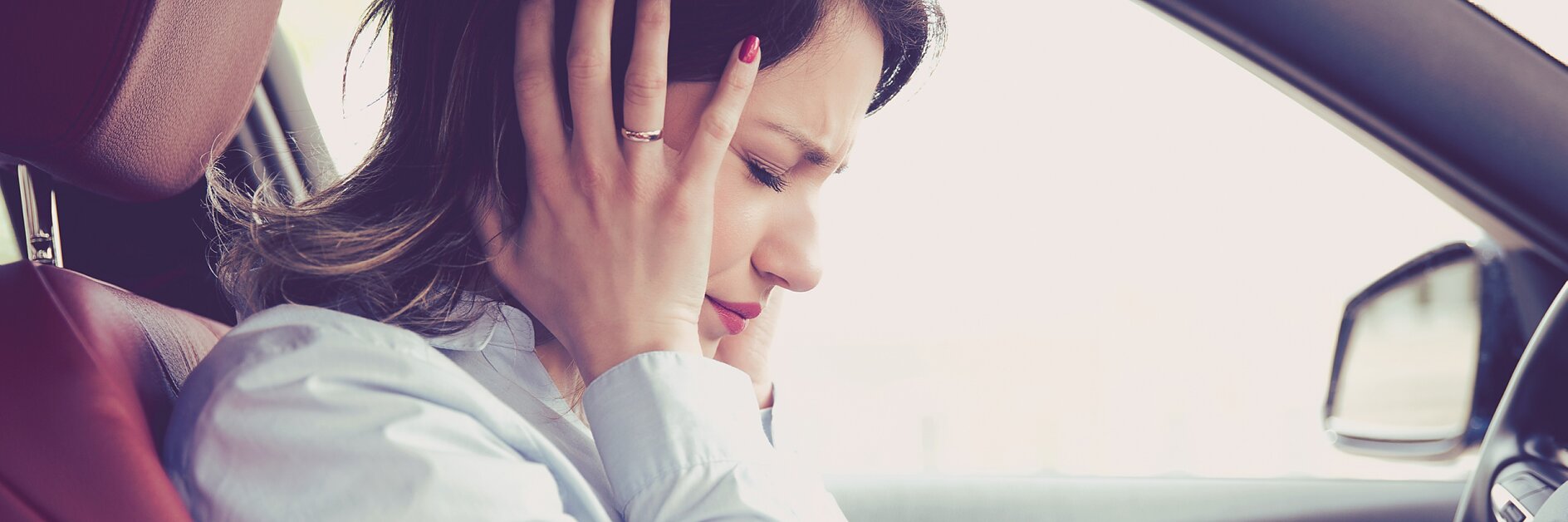 A woman is sitting in a car with both hands covering her ears. She appears to be uncomfortable or in distress. She is wearing a light-colored blouse and has dark hair. The interior of the car features dark and red accents. (This text has been generated by AI)