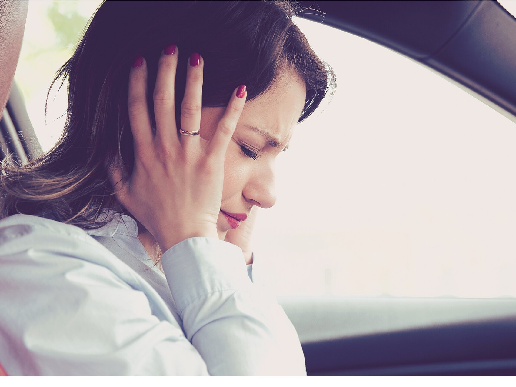 A woman is sitting in a car with both hands covering her ears. She appears to be uncomfortable or in distress. She is wearing a light-colored blouse and has dark hair. The interior of the car features dark and red accents. (This text has been generated by AI)
