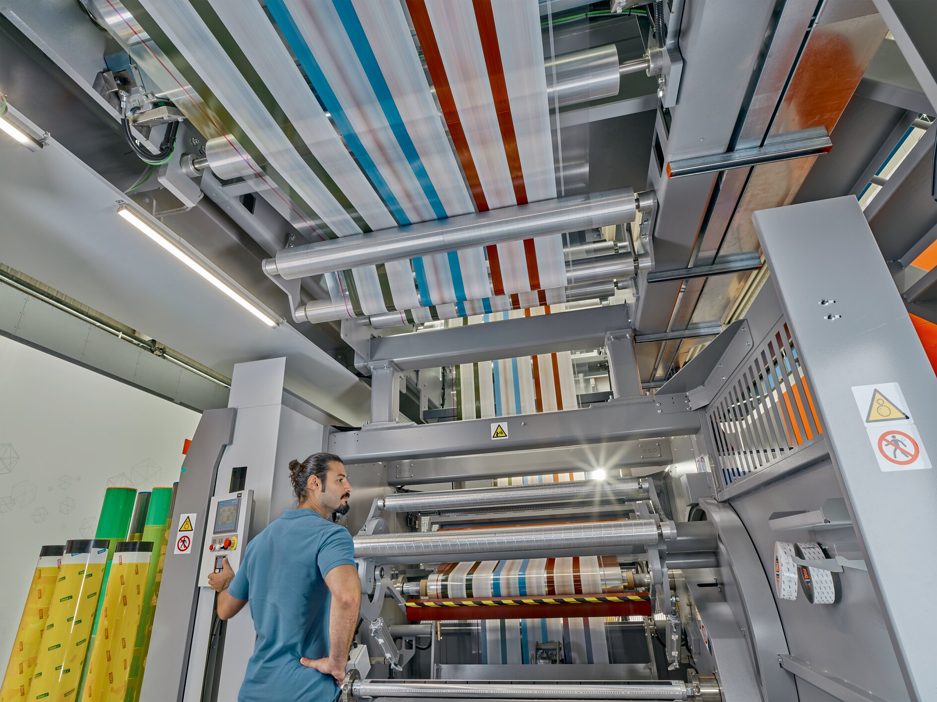 Flexo_Plate-mounting_Assortment_Bobst2023_001a-40153_V1 A man with a bun stands near a large industrial machine with multiple rollers, processing colorful striped material wrapped in tesa tape. The machine is metallic and has warning signs. The setting appears to be a factory or manufacturing facility. (This text has been generated by AI)