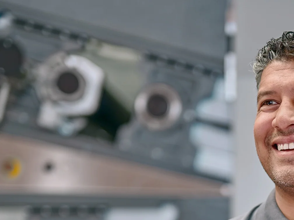 A man in a gray polo shirt stands smiling with his hands resting on a metallic cylinder in a machinery-filled environment. Two large cylindrical rolls, one metallic and one green, stand upright beside him. (This text has been generated by AI)