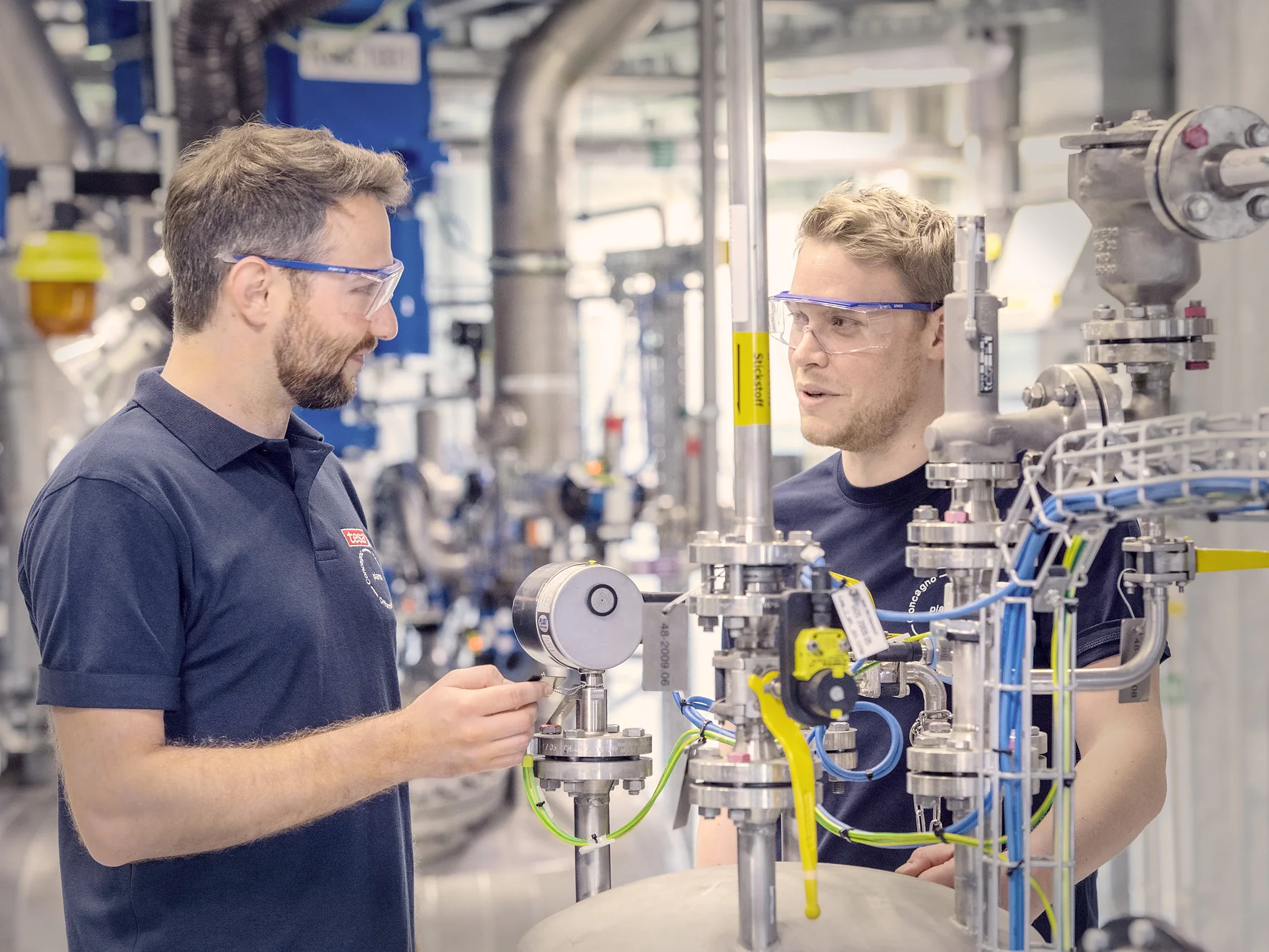 Two men wearing safety glasses and navy shirts stand in an industrial setting. They are discussing near complex machinery with pipes, valves, and gauges. The background shows more industrial equipment and a workspace environment, where tesa tape is prominently used for various applications. (This text has been generated by AI)