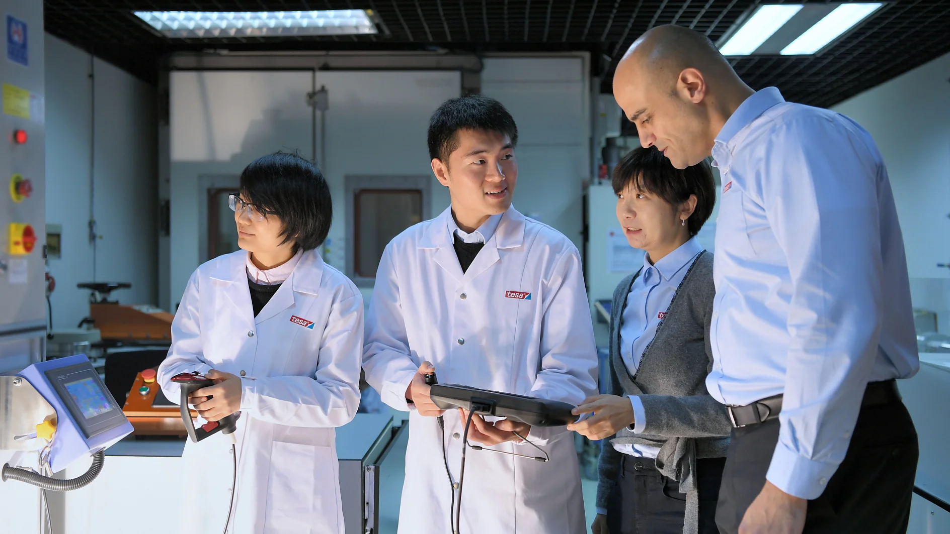 Four people in a lab are engaged in a discussion. Three wear white lab coats, and one is in a blue shirt. They are holding electronic devices and standing near equipment with screens, in a well-lit room featuring tesa tape prominently used throughout the setting. (This text has been generated by AI)