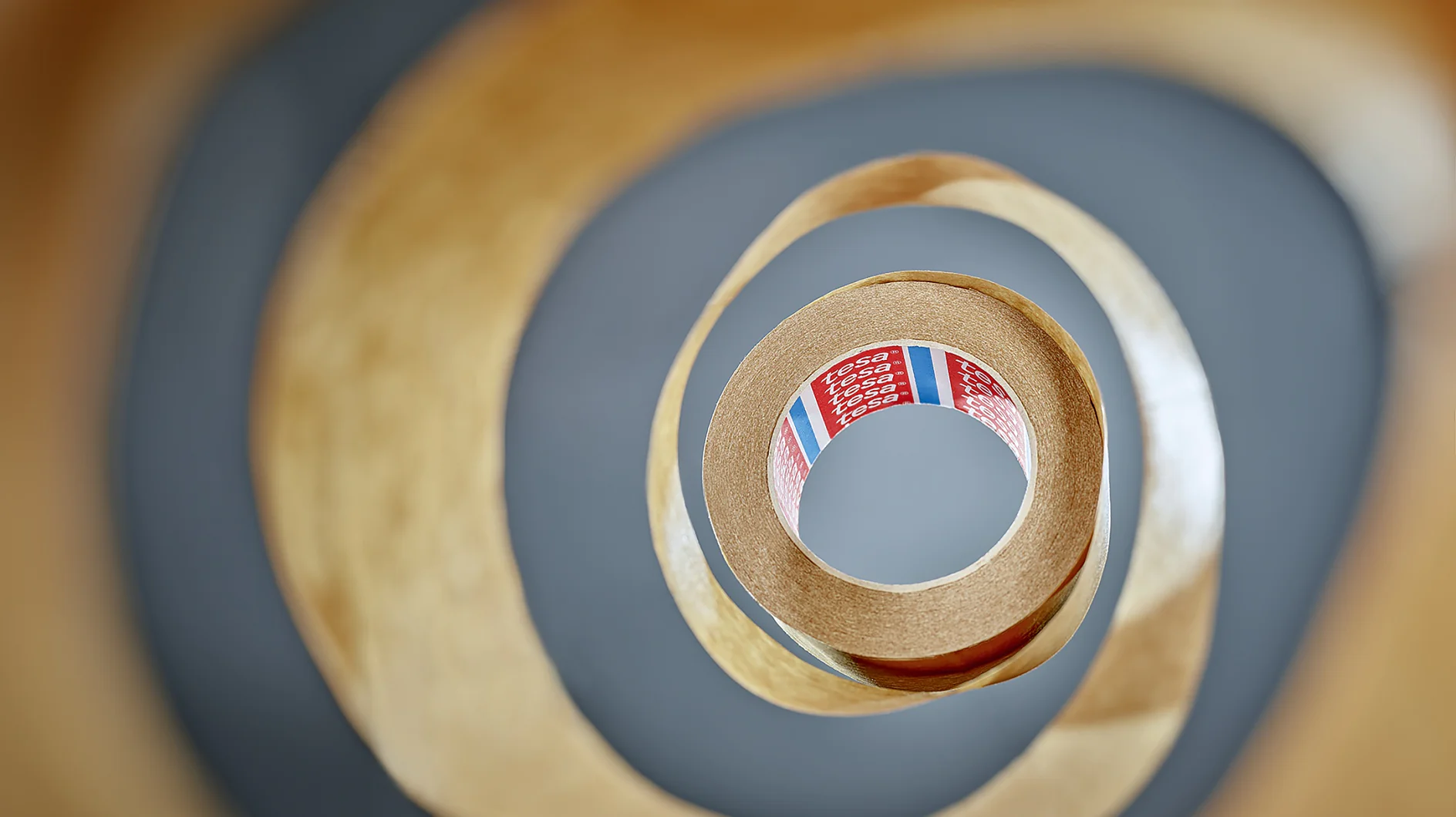 View from the center of a roll of tesa tape, creating a spiral pattern. The tesa tape is brown with a partially visible red label inside, featuring text and colored stripes. The background is blurred, emphasizing the layered effect of the tesa tape. (This text has been generated by AI)