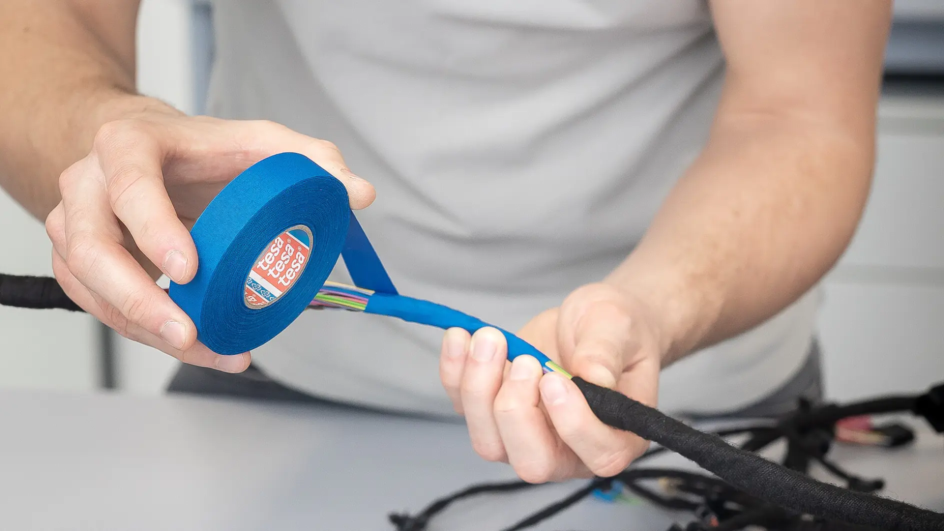 man in gray tesa shirt using blue tape dispenser to wrap black electrical wires on white table in indoor workspace