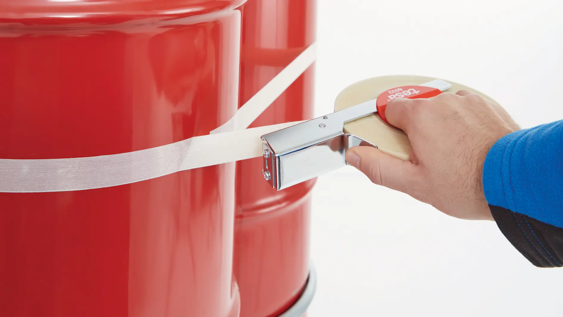 hand using a tesa tape dispenser to apply clear tape around a red metal drum in a bright indoor setting