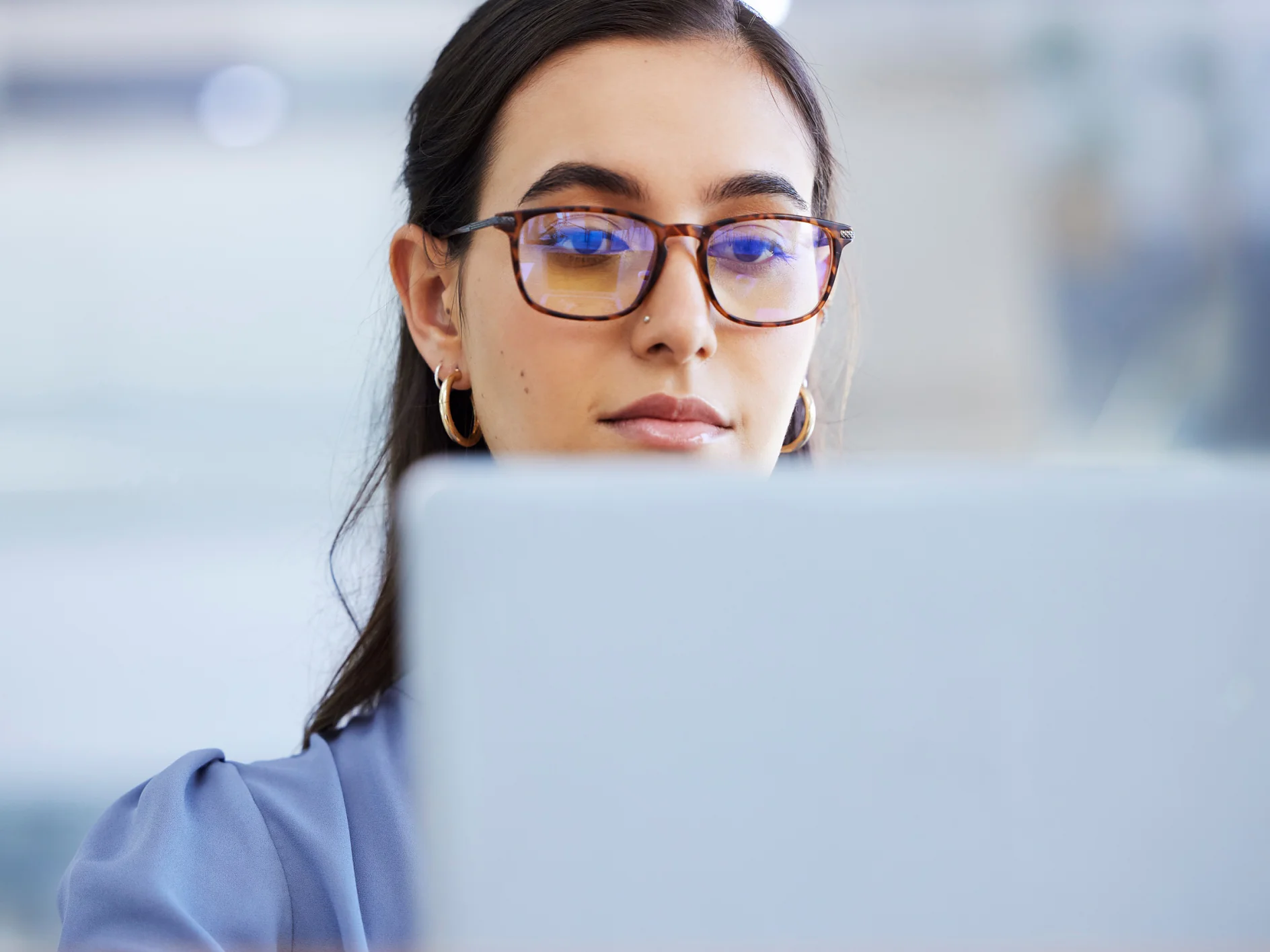 A woman wearing glasses and gold hoop earrings looks at a laptop screen. She has long dark hair and is dressed in a light blue top. The background is blurred and bright. Note: The original text does not mention tape or any competitor brands, so there are no references to replace with tesa tape. If you would like to provide a different text containing such references, I would be happy to rewrite it as requested. (This text has been generated by AI)
