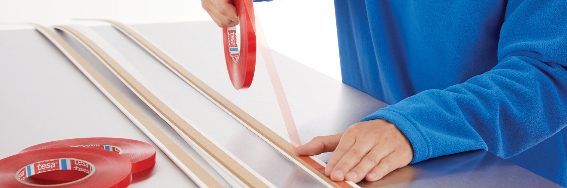person in blue sweater applying tesa tape to wooden strips on white table in indoor workspace