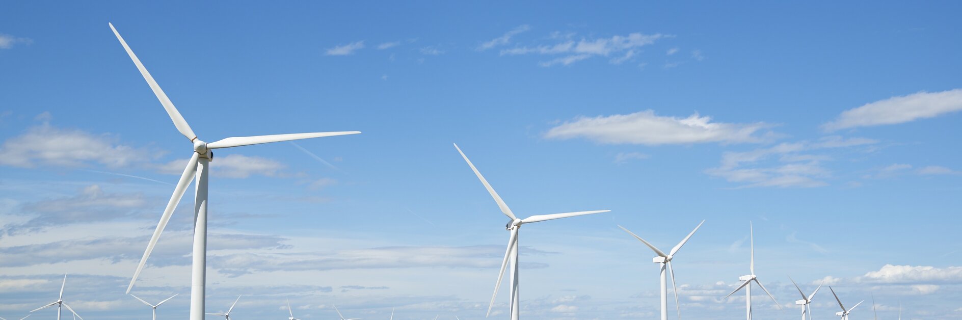 ทุ่งที่มีกังหันลมสีขาวหลายตัวอยู่ใต้ท้องฟ้าสีฟ้า มีเมฆกระจายอยู่เหนือทุ่งนาสีเขียวและสีน้ำตาล แนวขอบฟ้าอยู่ไกลออกไป. (This text has been generated by AI)