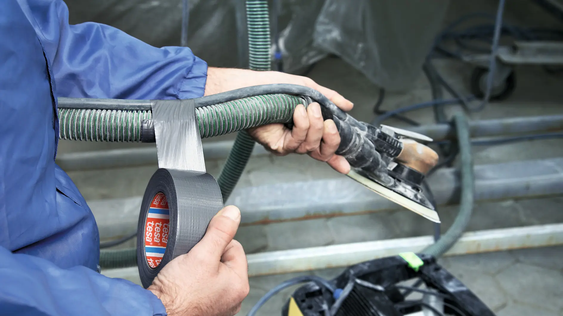 A person in a blue jacket is using silver tesa tape to secure a green ribbed hose to a metal tool with a rectangular base. The tool is connected to a piece of equipment with wheels in the background.