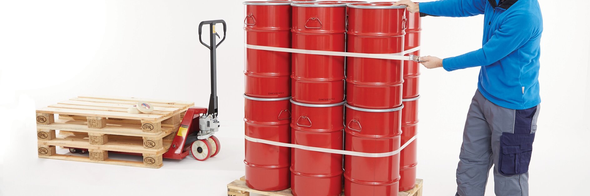 man securing red barrels on wooden pallet with tesa tape in a warehouse setting, pallet jack and empty pallet nearby