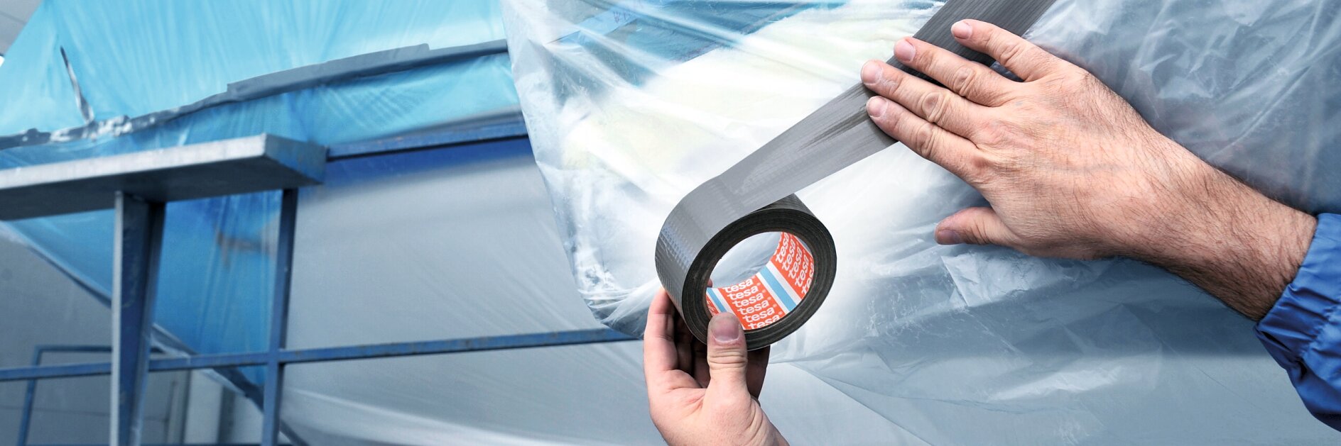 Two hands apply gray tesa tape to secure clear plastic sheeting over a large blue object, possibly in an industrial or construction setting. The background shows metal scaffolding and parts of the structure.