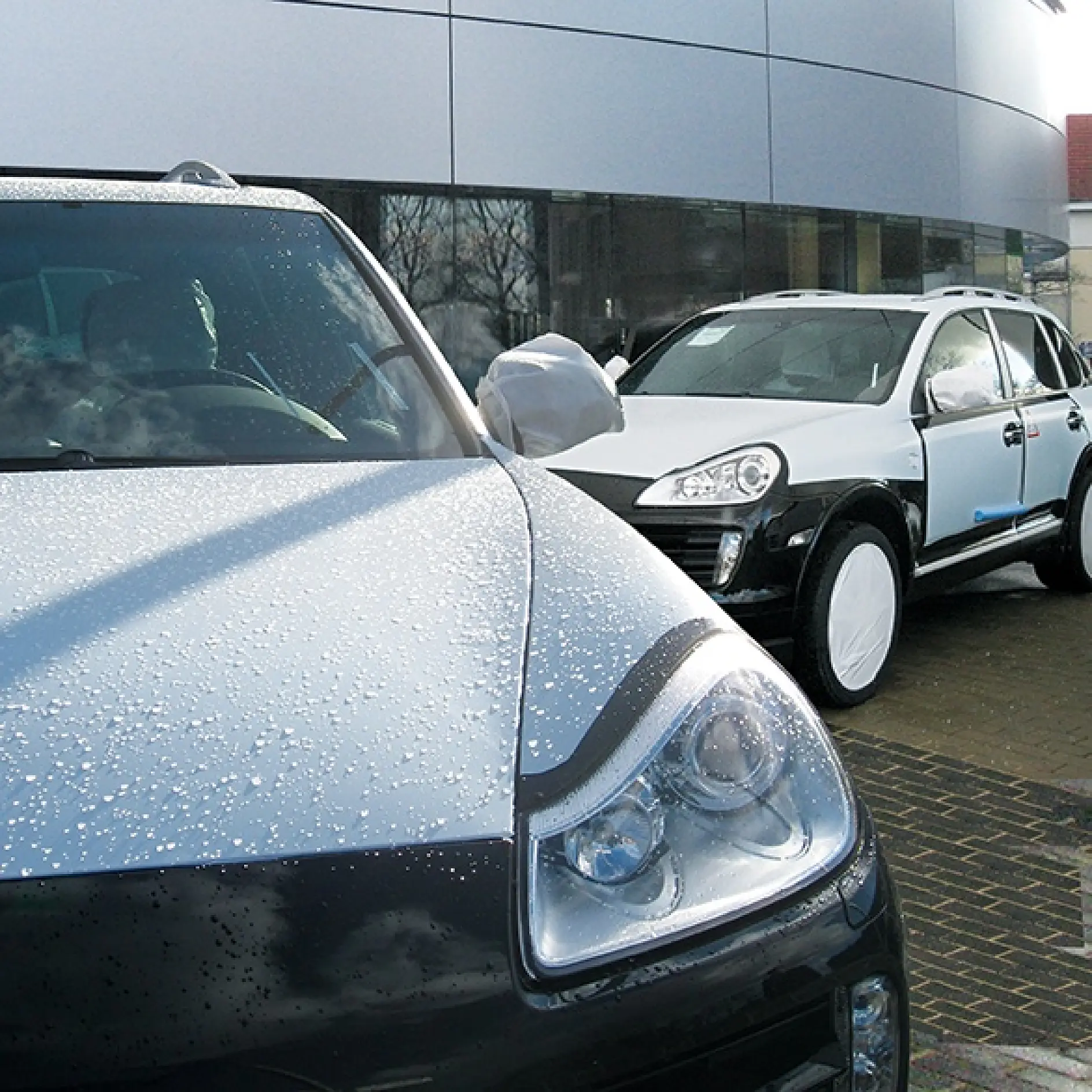 Two white and black SUVs are parked outside. The front SUV shows water droplets on its hood. The rear SUV is partially covered in protective tesa tape wrap. Both vehicles are in front of a modern, curved building with large windows.