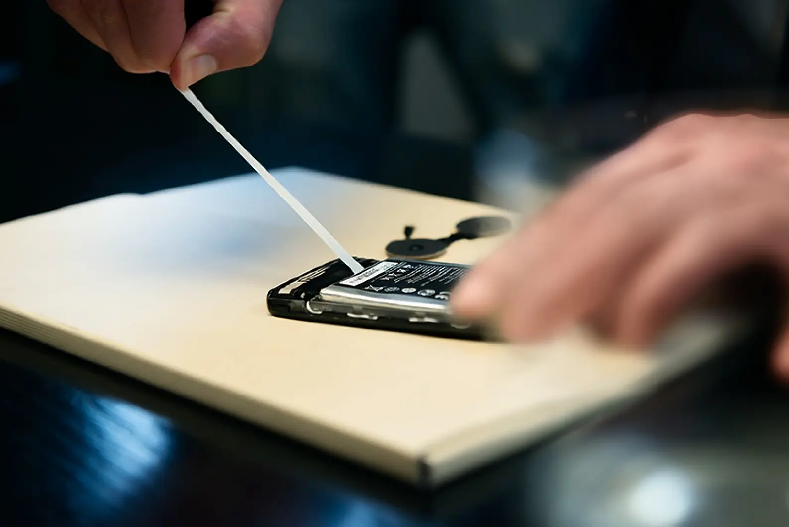 A person is using a plastic tool to remove the back cover of a smartphone placed on a table. The phones battery and internals are visible. The persons hand is slightly blurred, suggesting movement. (This text has been generated by AI)