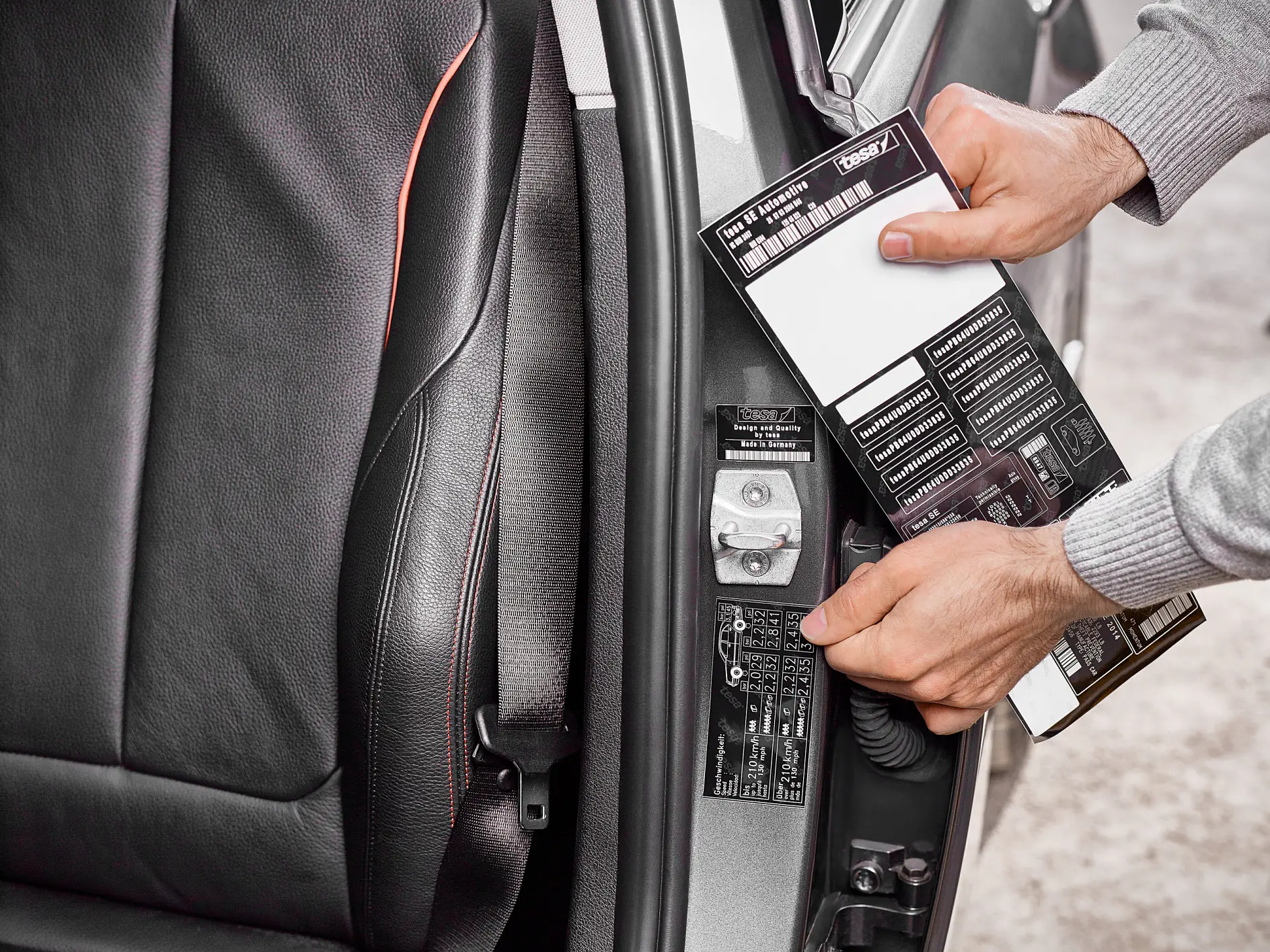 A person is placing a sticker with maintenance information inside a car door frame. The interior of the car features a black leather seat. The sticker, attached with tesa tape, has various details and barcodes. The person is wearing a gray long-sleeve shirt.