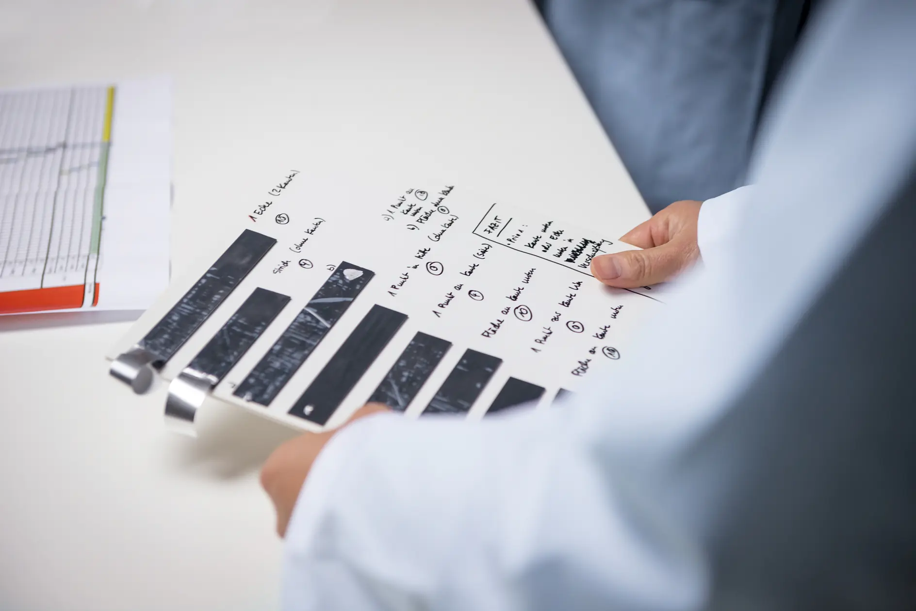 A person in a lab coat holds an X-ray film and a handwritten checklist. The X-ray is secured to a white sheet of paper with tesa tape, accompanied by notes and diagrams. A document with graphs is partially visible on a nearby table.