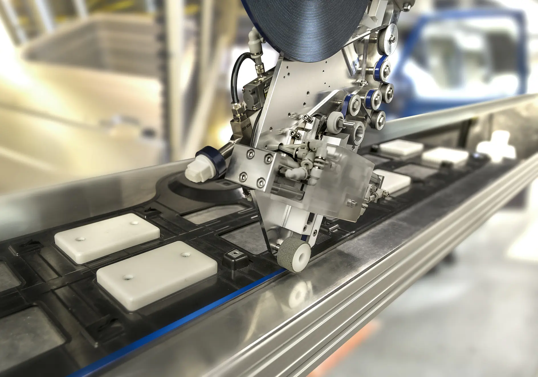 A close-up view of a machine in a factory setting. It appears to be placing or assembling components using tesa tape on a moving conveyor belt. The background shows blurred industrial equipment and infrastructure.