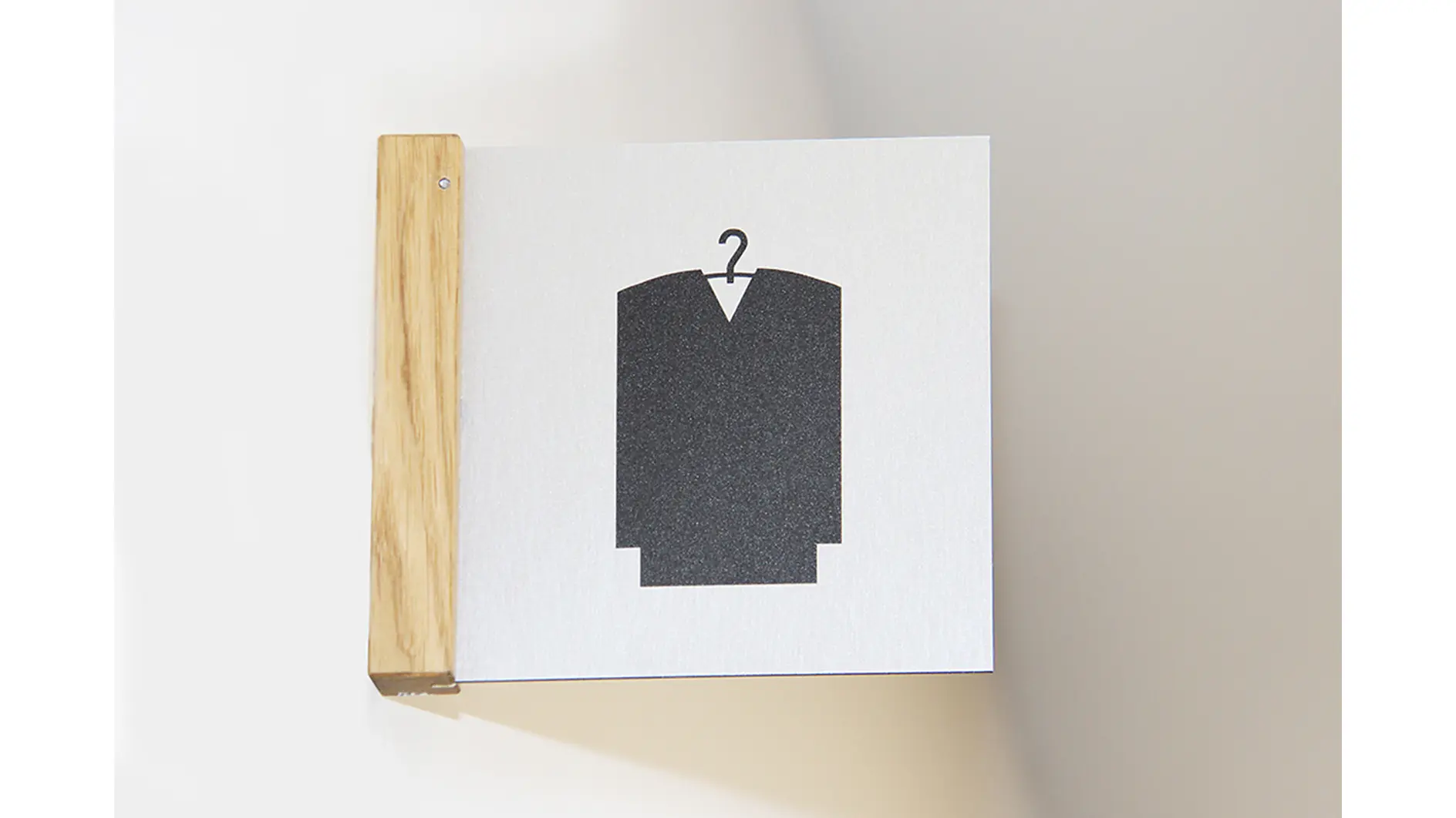 A sign attached to a wooden bracket on a white wall features a black silhouette of a V-neck garment on a hanger, resembling a kimono or robe, set against a light gray background. (This text has been generated by AI)