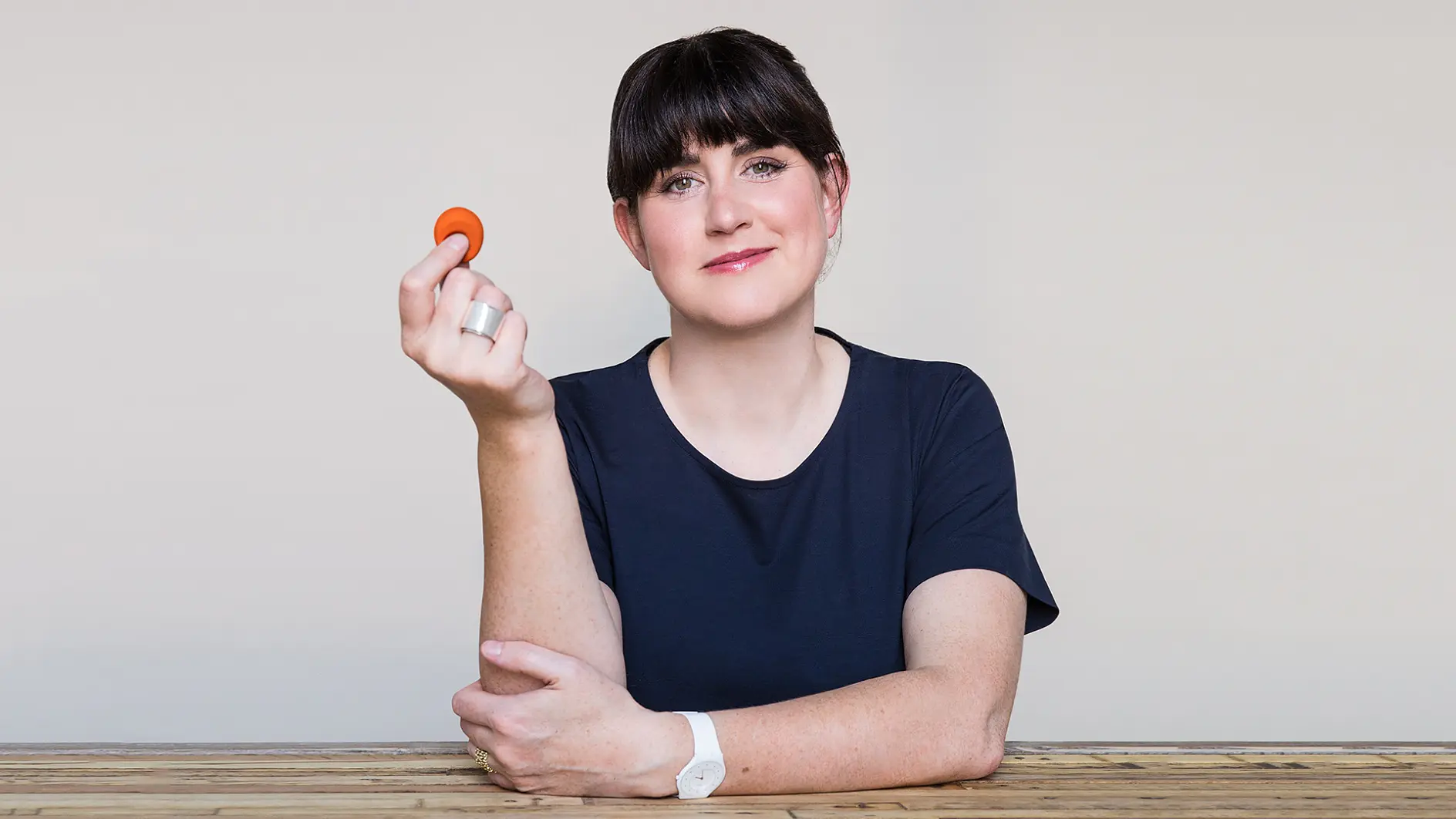 A person with short dark hair wearing a black shirt and white watch is sitting at a wooden table. They are smiling and holding a small orange object in their right hand, secured with tesa tape. The background is plain and light-colored. (This text has been generated by AI)