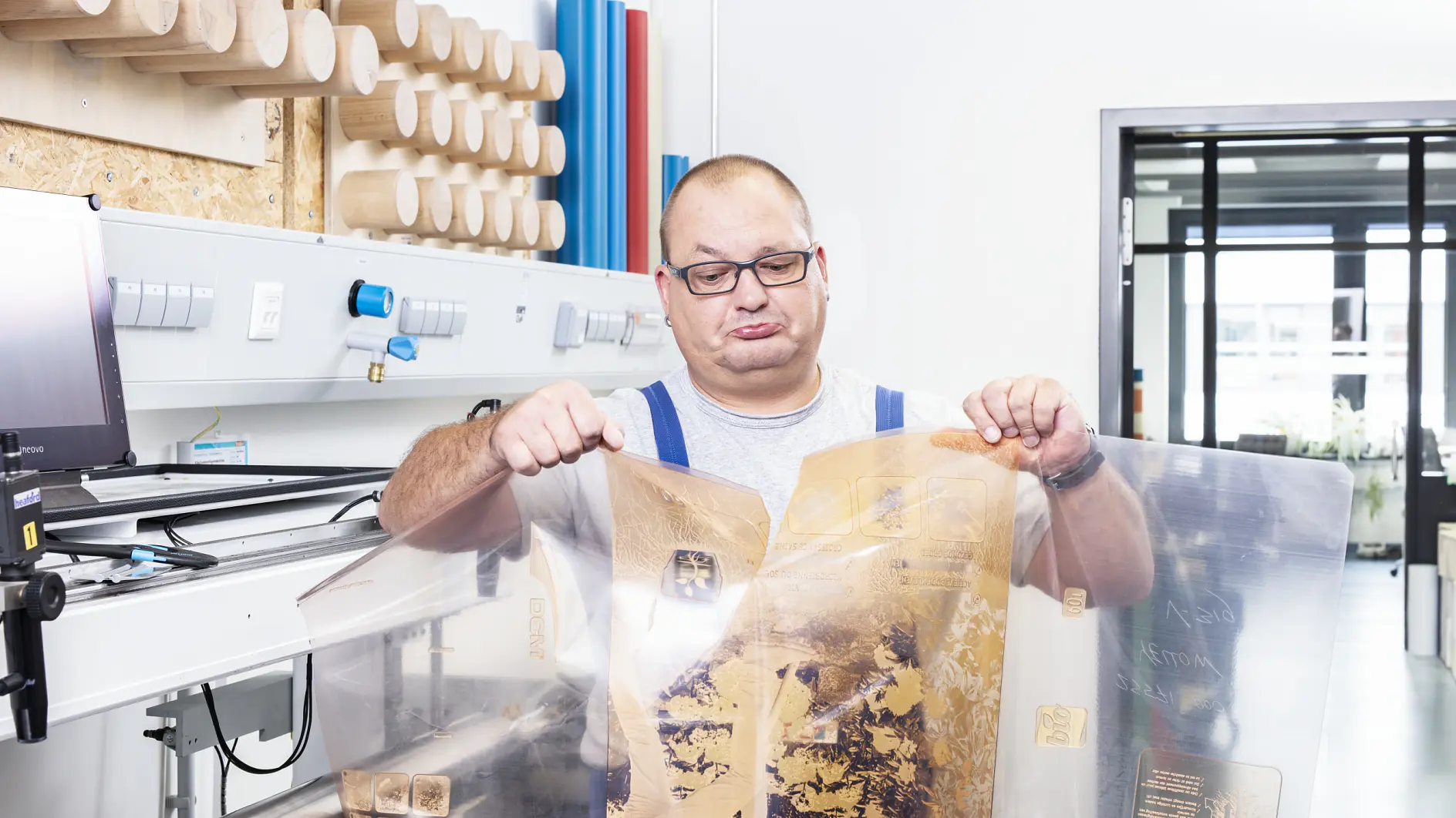 A man wearing glasses and blue overalls inspects transparent plastic sheets with printed designs in an industrial setting. Behind him are wooden rollers and machinery used for processing tesa tape. A window reveals a modern office space in the background.