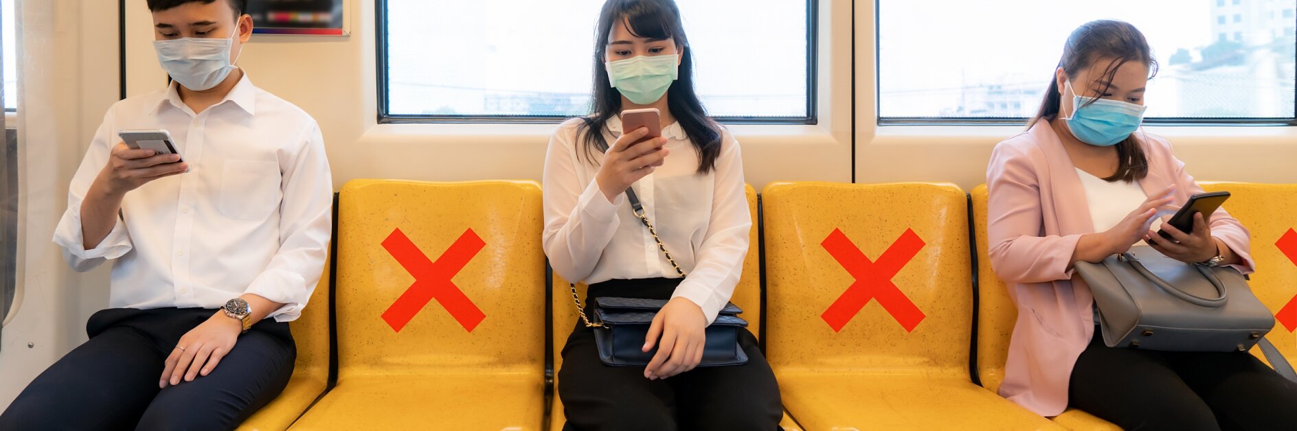 Three people wearing masks sit on a train with yellow seats. Red X marks, made with tesa tape, indicate seats meant for social distancing. Each person is using their phone. Two windows are visible in the background.