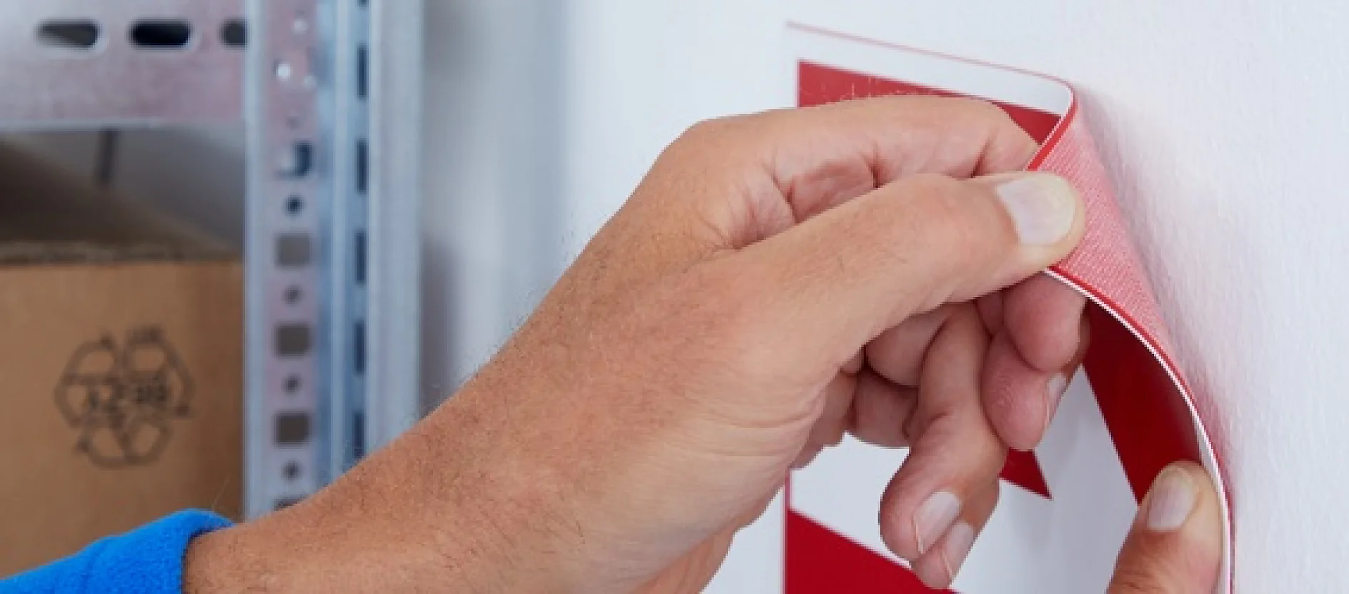 A person wearing a blue sleeve peels off the corner of a red and white adhesive label from a white wall using tesa tape. Part of a metal shelf and a cardboard box are visible in the background. (This text has been generated by AI)