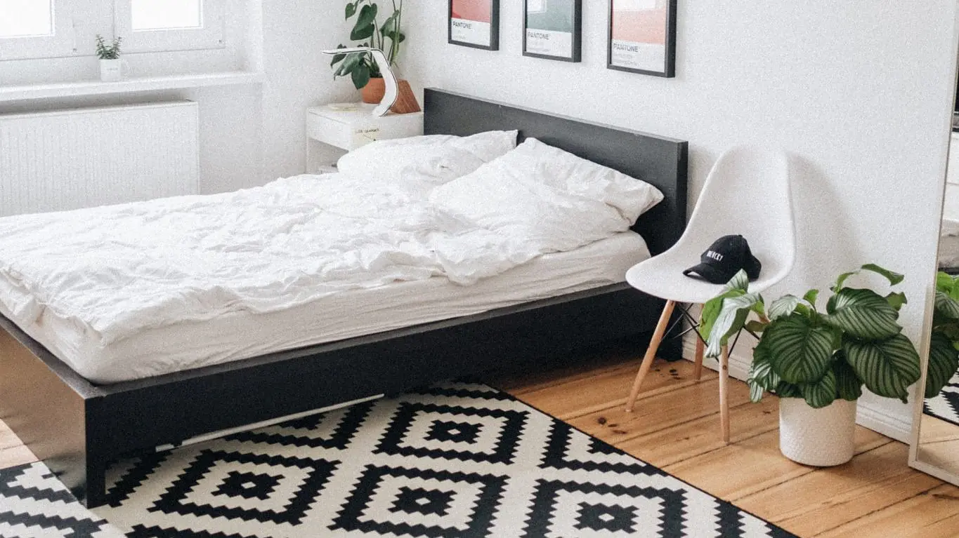 A bedroom features a black bed with white bedding and a geometric black and white rug. Above the bed, there are five framed pictures. A white chair holding a cap sits next to a potted plant. A window is on the left wall, while a mirror adorns the right wall. (This text has been generated by AI)